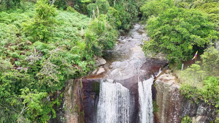 Drone pulls back from the top of a tiered waterfall in Colombia, revealing the full falls, surrounding tropical forest, and a small cabin near the cliff.