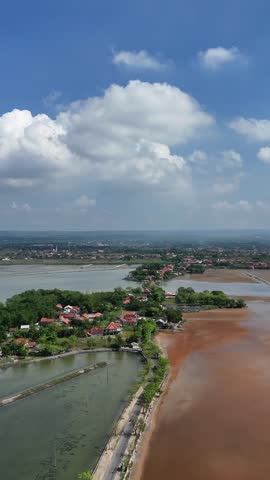 Vertical aerial view of a rural coastal village surrounded by salt pans and fish ponds under a blue cloudy sky in Indonesia