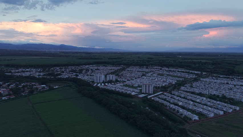 Drone video showcasing a scenic aerial view of a residential area in Valle del Cauca, Colombia, at sunset. Lush greenery surrounds neatly arranged homes under a colorful evening sky.