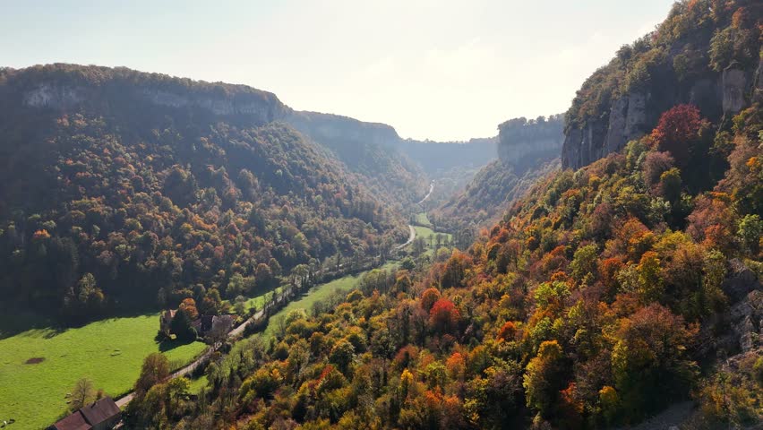 Aerial view of Baume les Messieurs in France featuring a winding river running through a valley surrounded by dense autumn forests, dramatic limestone cliffs, and rural fields nestled within landscape