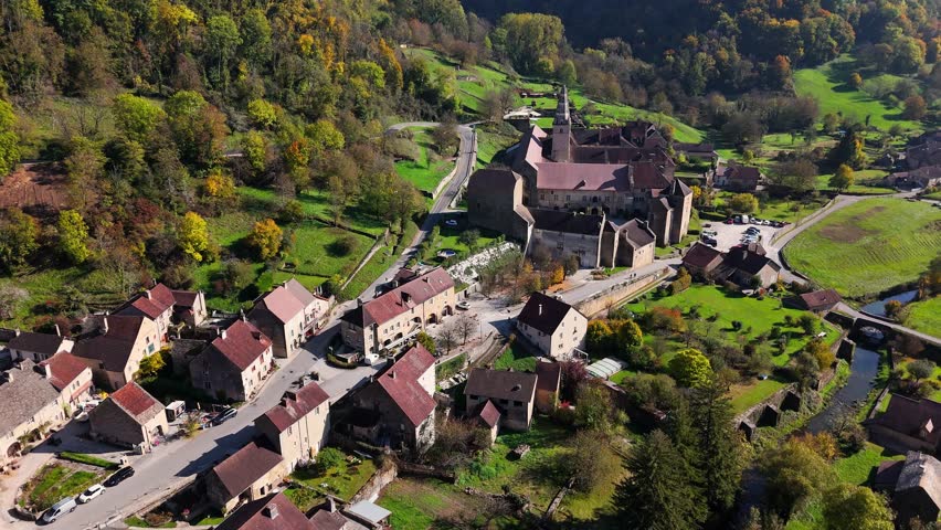 Aerial perspective of Baume les Messieurs in France featuring a medieval village, historic abbey, clustered houses, winding roads, and lush countryside framed by forests, fields, and rolling hills