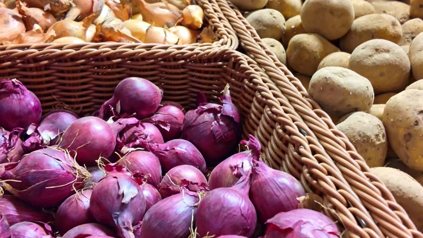 Descending shot along a market stall showing potatoes and onions before revealing red onions in a wicker basket.