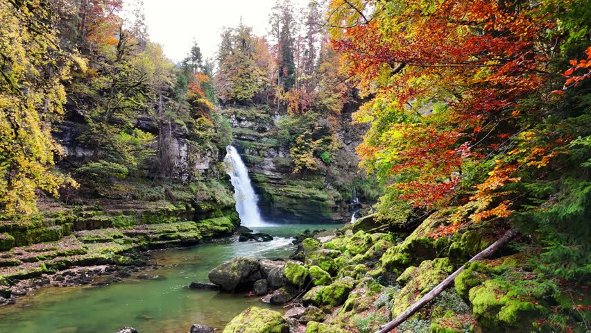 Saut du Doubs waterfall in France with clear green water, moss covered rocks and vibrant autumn forest. Steep cliffs and colorful foliage frame the flowing river, creating a peaceful natural landscape