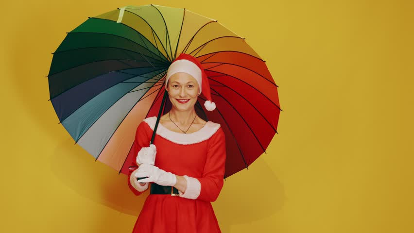 Smiling woman wearing a santa claus helper costume and hat holding a large colorful rainbow umbrella while posing on a yellow background
