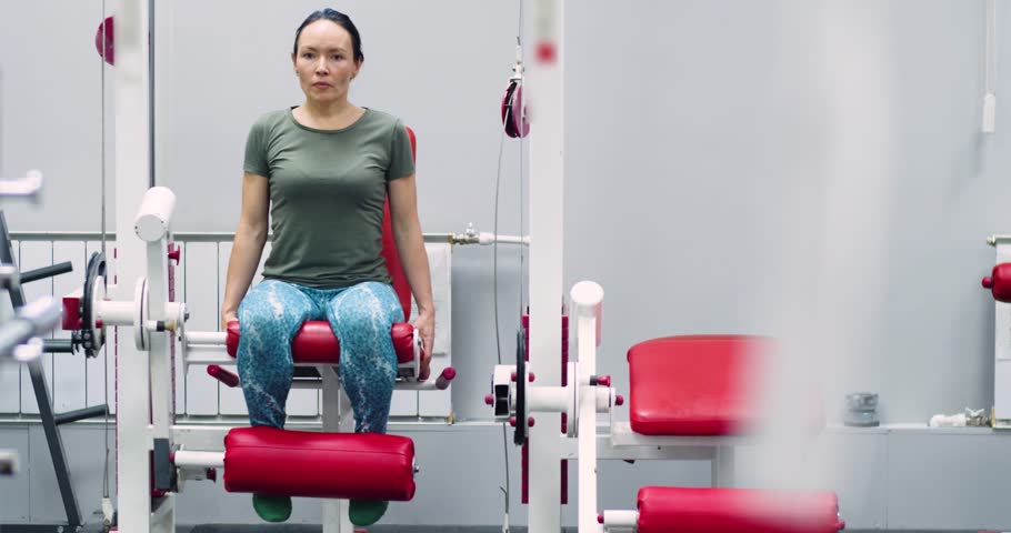 Slow motion 4k footage of a middle-aged woman during gym workout. Leg extensions in gym fitness exercise.