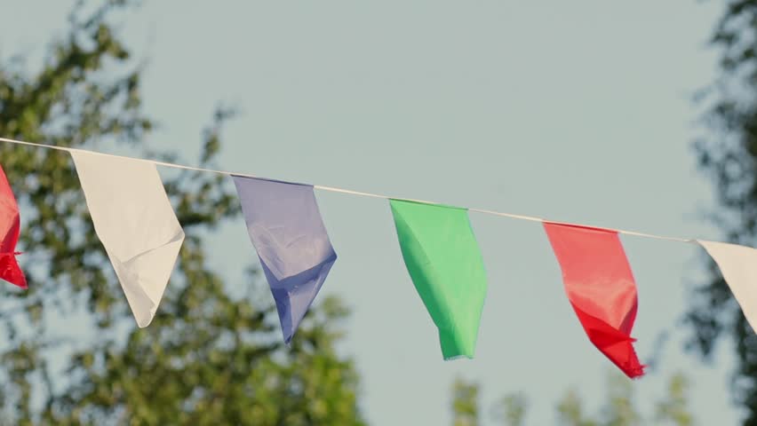 A row of colorful pennant flags flutters gently on a string against a clear sky, with softly blurred green foliage in the background, creating a light and festive outdoor atmosphere.