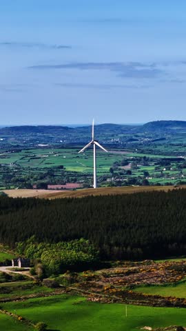 The wind turbines are situated on a hilltop overlooking a lush green forest. In the distance, green fields and mountains stretch to the horizon under a bright blue sky.
