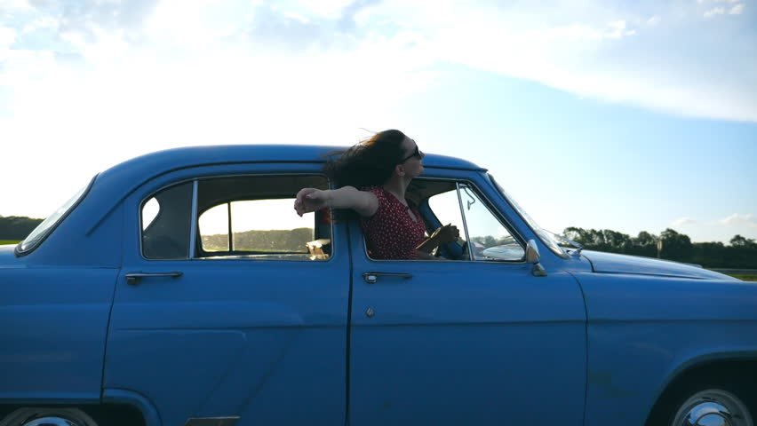 Young attractive girl in sunglasses leaning out of vintage car window and enjoying trip in summer time. Woman looks out from moving retro vehicle. Travel concept. Side view Slow motion