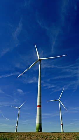 Vertical video of windmills against a blue sky. Wind turbines spinning and generating energy in a field. Alternative energy production.