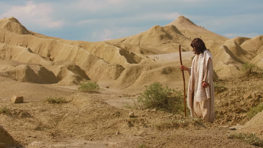Representation of Christ in ancient-style clothing walks through a barren desert landscape, surrounded by sculpted sand formations and warm golden light. Cinematic scene evoking solitude, spirituality and timeless exploration. Christian concept. Slow motion footage