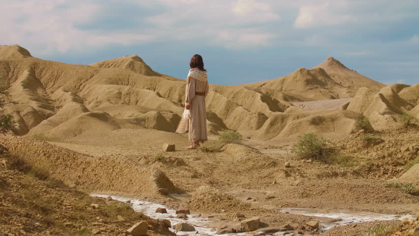 Wide Shot of a human representation of Christ in traditional biblical-style clothing, walking through an arid desert landscape, surrounded by sculpted sand formations and warm golden light. Cinematic scene evoking solitude, spirituality and timeless exploration. Christian concept. Slow motion shot