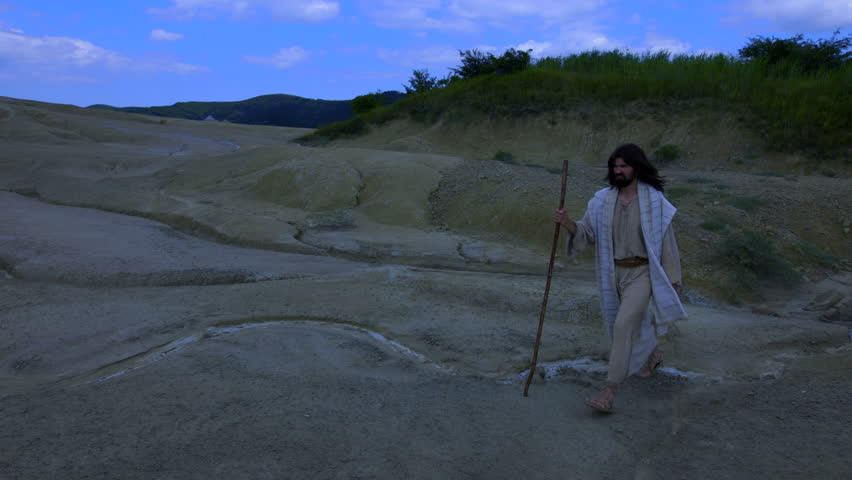 Representation of Christ, wearing ancient robe, walks away into the night, crossing a barren and rugged landscape. A dry white stream runs through the foreground, against a dramatic blue sky, emphasizing the isolation of the spiritual journey.