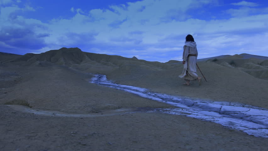 The solitary figure of the Christ walks away into the night, crossing a barren and rugged landscape. A dry white stream runs through the foreground, against a dramatic blue sky, emphasizing the isolation of the spiritual journey. Super slow motion shot