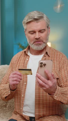 Caucasian mature man sitting on home sofa, holding smartphone and credit card, making money transfer through mobile app. Middle-aged guy looks satisfied with results, pointing finger at smartphone
