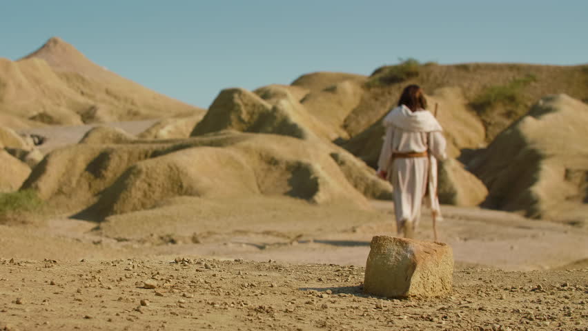 Long Shot of a Solitary Traveler in simple traditional attire crosses an arid, sunlit terrain, using a wooden staff while walking through a quiet and rugged wilderness. Christian and spiritual concept. Slow motion footage