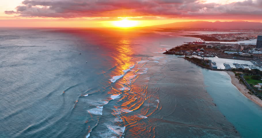 Mesmerizing setting sun colors the sky in orange, the heavy cloudscape and the waves of the Pacific slowly rolling to the coast. Shore of Honolulu, Hawaii at sunset. Aerial perspective.