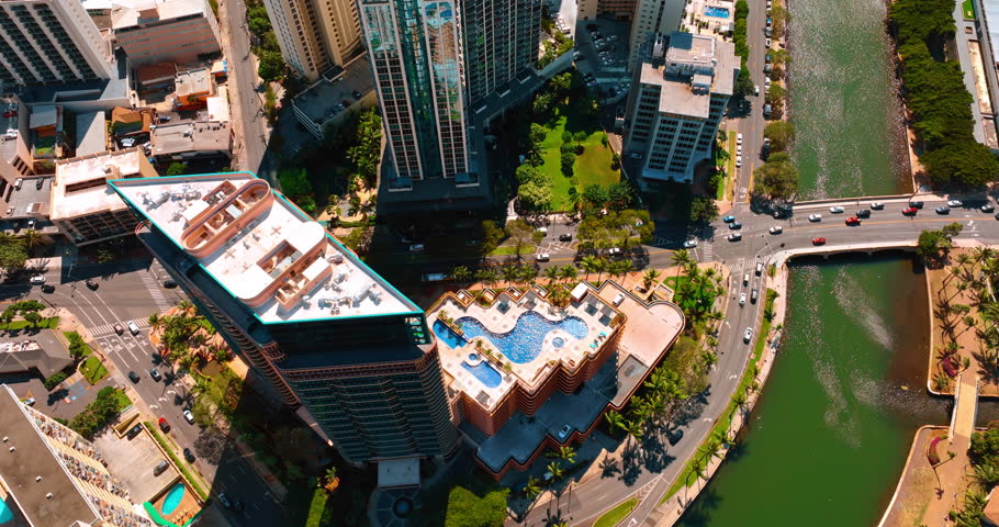 Lots of cars move by the modern roads and bridge crossing the Ala Wai Canal. Drone footage over the building with a swimming pool on the roof. Waikiki resort in Honolulu, Hawaii, USA.
