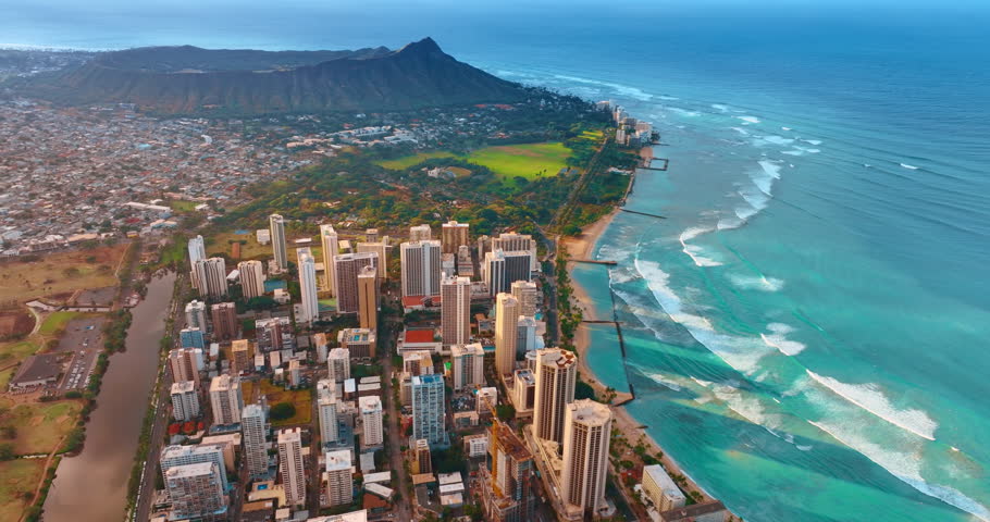 White foamy waves roll slowly to the iconic sandy beaches. Drone footage over the cityscape of Waikiki in Honolulu, Hawaii, USA. The Diamond Head Crater and backdrop.