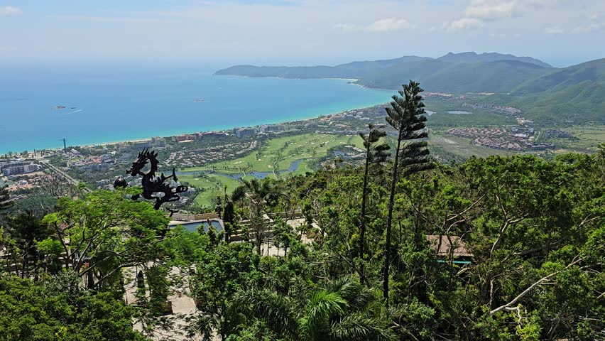 Aerial view of Yalong Bay beach in Sanya, China. Turquoise ocean, white sand coastline, green mountains and resort hotels. Tropical paradise with dragon statue landmark and golf course.
