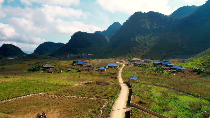 Aerial tracking shot flying along a narrow, winding road leading into a colorful Hmong ethnic village nestled in the mountains of Hang Kia Pa Co, Mai Chau, Vietnam, illuminated by golden hour sun.