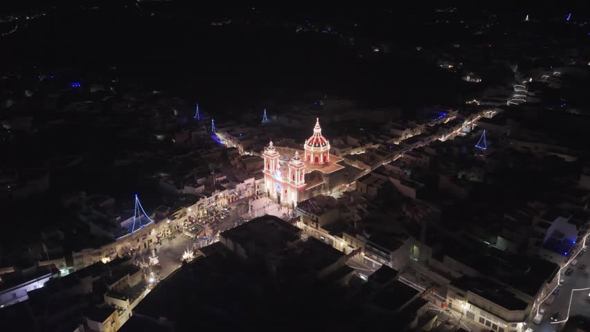 Aerial night view of the illuminated church in Xagħra, Gozo, showcasing its bright façade and surrounding streets lit up against the dark urban landscape.