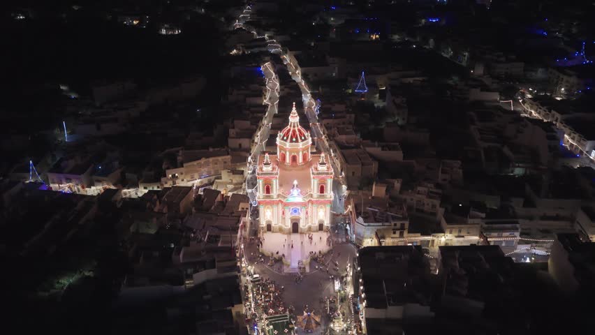 Aerial 6K night view of the illuminated church in Xagħra, Gozo, showcasing its bright façade and surrounding streets lit up against the dark urban landscape.
