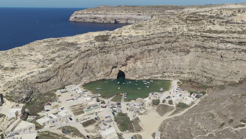 Aerial 6K view of the Inland Sea in Dwejra, Gozo, showing the natural stone arch tunnel, turquoise water, cliffs and the small fishing village.
