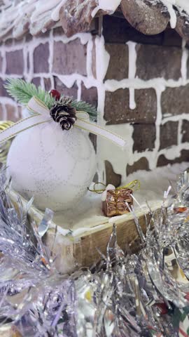 Closeup vertical video of Christmas décor featuring a white glitter bauble, silver tinsel garland, a gingerbread-style snow brick house, and a tiny gift box in a festive winter setting.