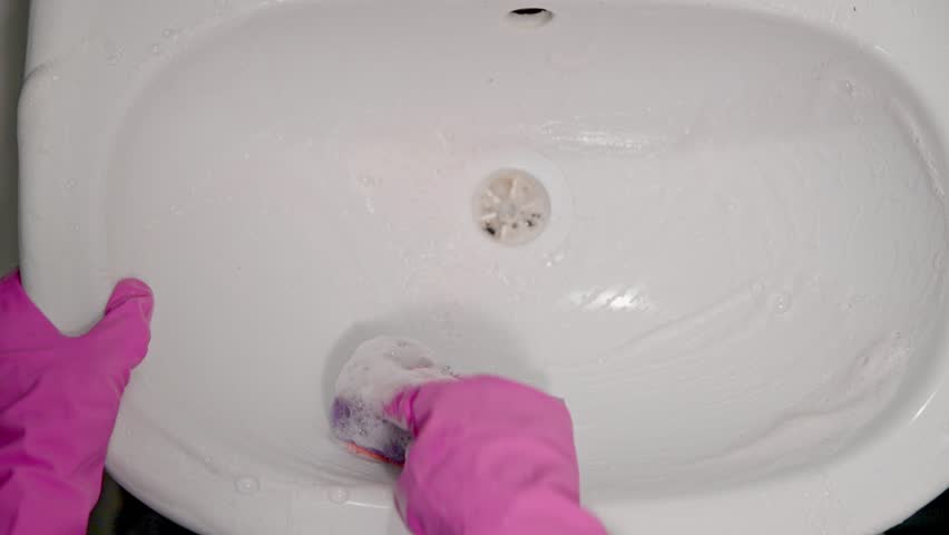 Person wearing pink rubber gloves scrubs white sink surface actively, with visible cleaning motion and soap suds. Bright lighting highlights cleanliness and hygiene effort