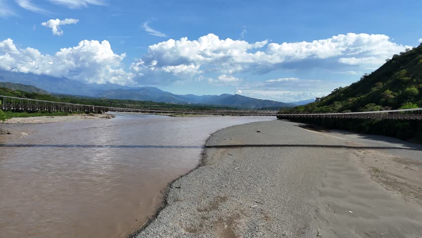 An immersive POV shot approaching the Puente de Occidente (Western Bridge), a historic 19th-century suspension bridge over the wide Cauca River in rural Antioquia, Colombia