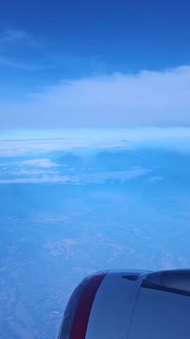 Vertical aerial view from an airplane window showcasing distant, hazy mountain peaks and a vast landscape below, with the aircraft engine partially visible