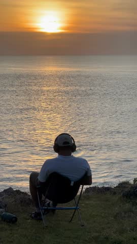 Rear view of a man on a cliff wearing headphones, enjoying the beautiful sunset over the ocean. Concept for relaxation, music, and travel escape.