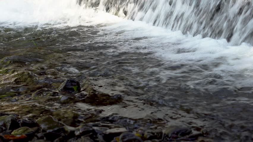 A low-angle shot showing a waterfall or small rapids falling into a river below. The waterfall creates white foam, while the foreground water ripples and shimmers over small rocks on the riverbed.