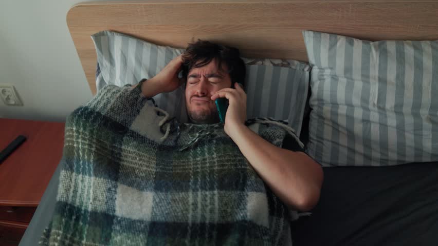 A man lies in bed under a blanket while talking on the phone, showing a painful, stressed expression. A realistic morning moment capturing discomfort and emotional tension.