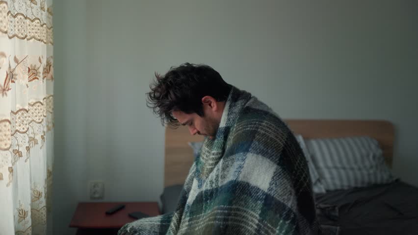 An exhausted man sits on the edge of the bed wrapped in a blanket, struggling with low morning energy. His tired posture reflects fatigue and reluctance to start the day.