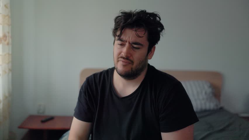 A weary man sits on the edge of his bed wrapped in a blanket, looking tired as he checks his phone. His expression reflects low energy and the difficulty of starting the day.
