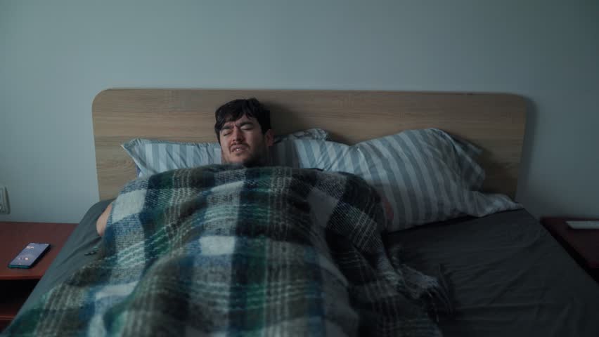 A sleepy man struggles to get out of bed in the morning, wrapped in a blanket and showing a strained, fatigued expression as he begins his slow daily wake-up routine.