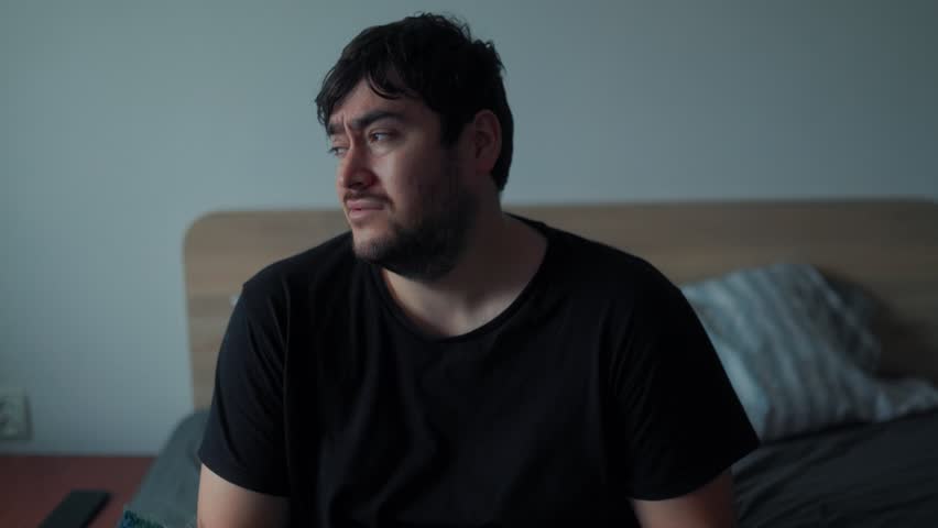 A tired man wakes up slowly in the morning, looking groggy and uncomfortable as he reacts to the early hour and struggles to start his day, wrapped in a warm blanket.