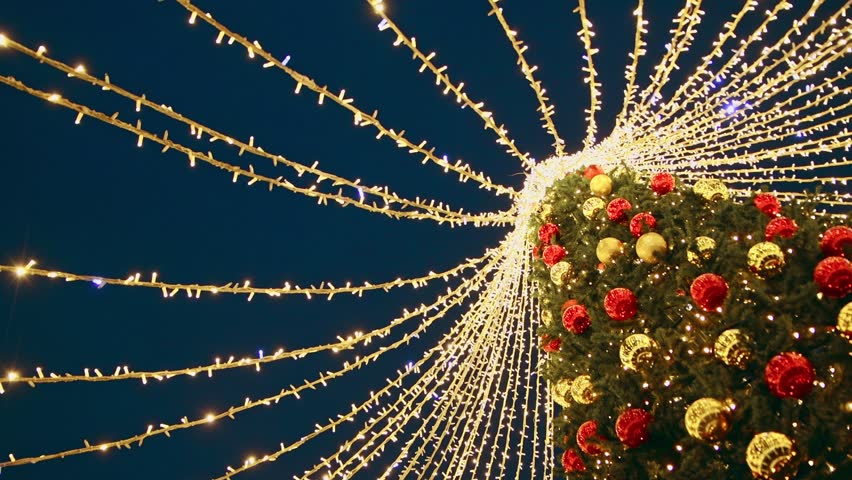 Tall christmas tree decorated with red and gold ornaments features cascading warm white string lights radiating outward under dark night sky, creating festive and vibrant holiday atmosphere
