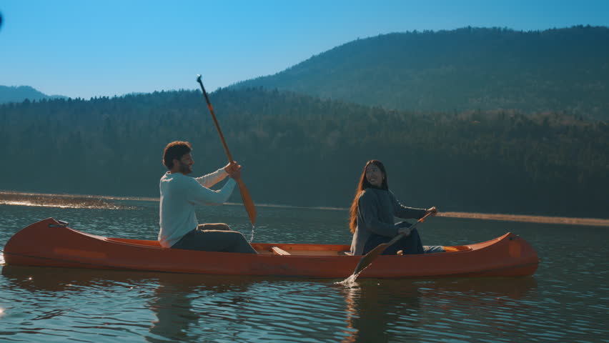 A couple paddles a canoe on a serene lake, surrounded by forested hills. They enjoy a peaceful weekend activity, embracing, captured with a full shot of subject, an active lifestyle.