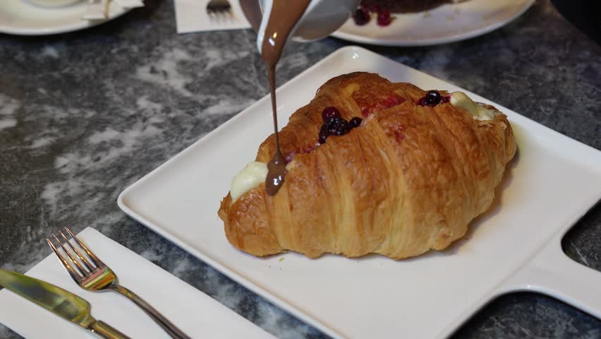 Chef drizzling liquid chocolate topping over a golden croissant pastry.