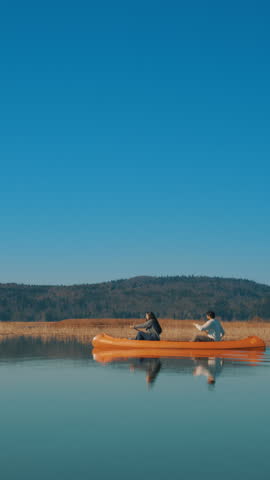 A couple paddles a canoe on a serene lake, surrounded by a clear blue sky and distant hills. The calm water reflects their peaceful activity, emphasizing tranquility and connection in nature.