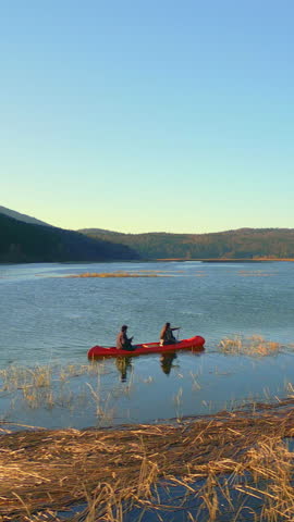Two individuals paddle a red canoe on a serene lake, surrounded by golden reeds and distant hills. The scene captures a peaceful weekend activity, emphasizing connection with nature.