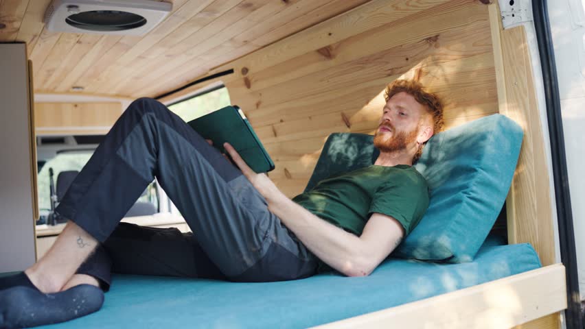 Young redhead man relaxing on the bed inside a modern camper van. His focus is on reading an e-book on a tablet during his road trip vacation