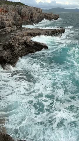 Powerful turquoise ocean waves crash against the rugged rocky shore. Turbulent seawater laps against the rocks during a storm