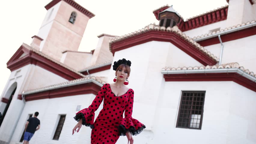 Beautiful woman in a traditional red polka dot dress dancing flamenco. Graceful and passionate performance in front of a white building in spain