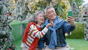 In a cozy outdoor setting, an elderly asian couple shares joyful moments beneath a beautifully decorated Christmas tree, snapping selfies and admiring their cheerful photos together in the winter - Powered by Shutterstock - Get 15% off with code: PIKWIZARD15