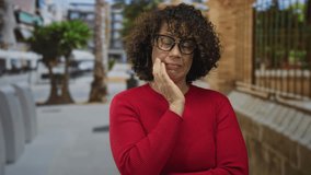 Woman with hand on chin on urban street beside brick building facade in casual red sweater and glasses; doubt. - Powered by Shutterstock - Get 15% off with code: PIKWIZARD15