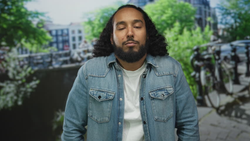 Man with beard stands on urban street looking at camera in denim jacket and white tee; quiet contemplative reflection.
