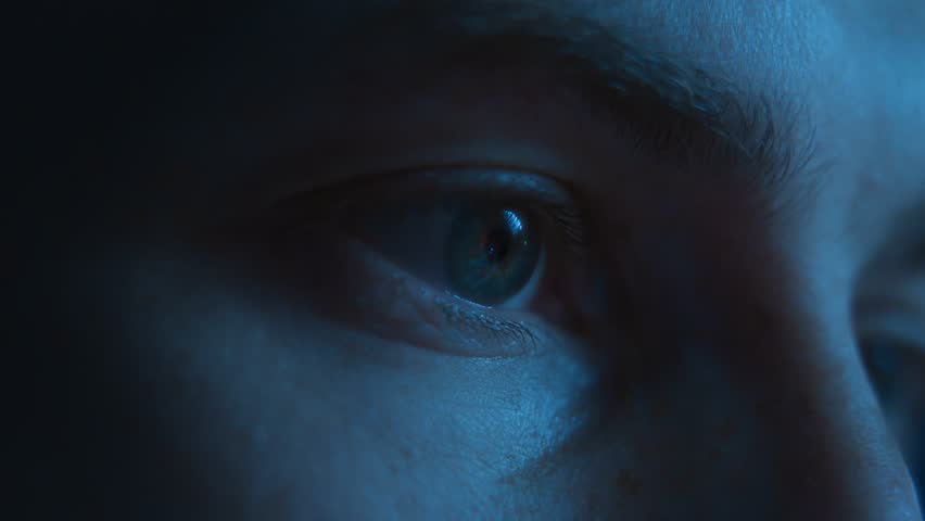 Close-up of man eyes looking with focus on a computer or smartphone display with the screen reflection in his pupil, working late at night or scrolling through a news feed, illuminated with blue light